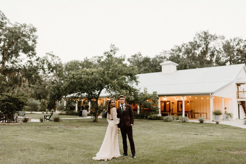 bride and groom standing together in grass outside of white barn with market lights